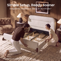 Two people kneel on a rug while positioning the cream upholstered sides of a partially assembled bed frame with exposed support rails and a tall rounded headboard.