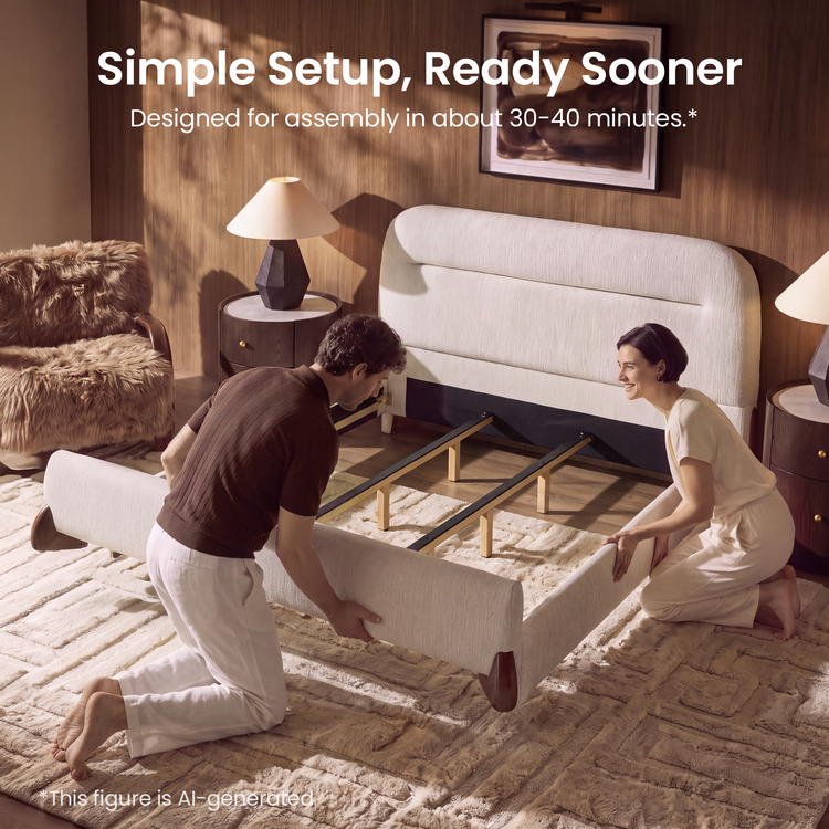 Two people kneel on a rug while positioning the cream upholstered sides of a partially assembled bed frame with exposed support rails and a tall rounded headboard.