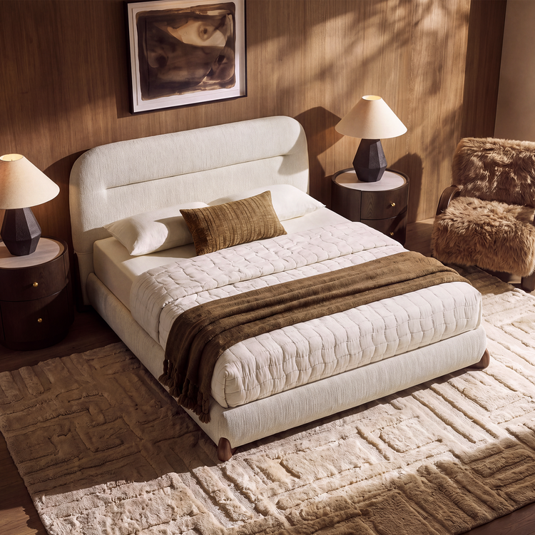 Cream upholstered bed with white quilted bedding and a brown throw, flanked by round bedside tables and lamps, with a shaggy brown chair and geometric cream rug.