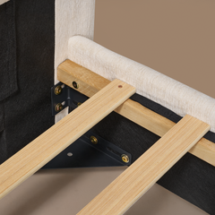 Close-up of an upholstered frame corner with pale slats, black brackets and gold-colored fasteners beneath cream textured upholstery.