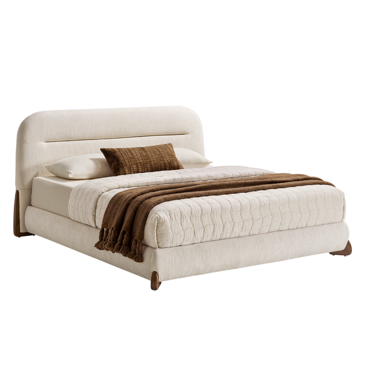 Cream upholstered bed with a rounded rectangular headboard and brown feet, dressed in cream quilted bedding, white pillows, a brown accent cushion and a brown tasseled throw.
