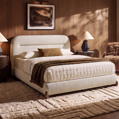 Cream upholstered bed with a rounded headboard, white quilt and brown throw, flanked by bedside lamps beneath abstract artwork in a sunlit bedroom.
