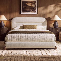 Cream upholstered bed with a rounded headboard, white quilt and brown throw, flanked by dark bedside tables and lamps beneath framed abstract art.
