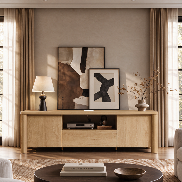 A light wood console stands beneath two abstract framed artworks, with a table lamp, a vase of branches, and audio equipment in a sunlit living room.