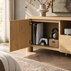 Open wooden media cabinet displaying a vertical game console, controller, headphones on a woven basket, and a silver electronic device, with a vase of dried branches on top.