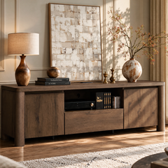 A dark wood console cabinet with three doors and an open shelf sits beneath a large abstract artwork, decorated with a lamp, books, small ornaments, and a vase of branches.