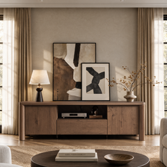 A brown cabinet holds a table lamp, two overlapping framed abstract artworks and a vase of branches, with audio equipment in an open shelf and tall curtained windows on either side.