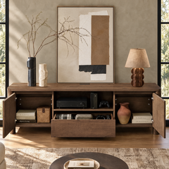 A wooden media cabinet with open doors and a central drawer holds electronics, books, and a vase; abstract framed art, two vases, and a table lamp sit on top.