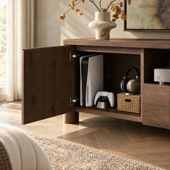 An open dark wood cabinet holds a white game console, controller, headphones, and a silver electronic device, with a vase and branches on top.