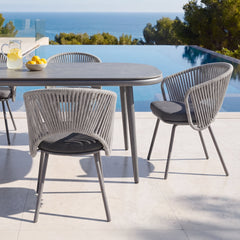 Gray rectangular outdoor dining table with four rope-back chairs beside a pool and ocean view
