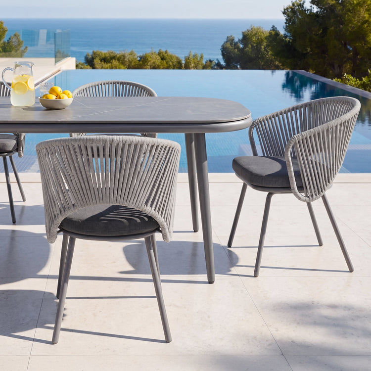 Gray rectangular outdoor dining table with four rope-back chairs beside a pool and ocean view