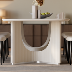 A light-colored dining table with fluted panels and a curved central opening stands between upholstered chairs, with a bowl of green fruit and vases on top.