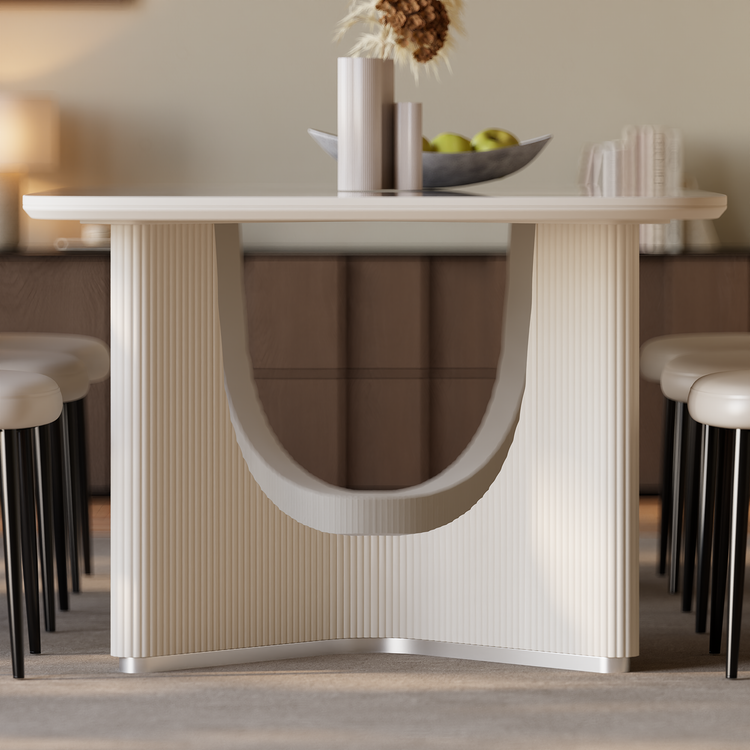 A light-colored dining table with fluted panels and a curved central opening stands between upholstered chairs, with a bowl of green fruit and vases on top.