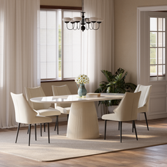 Bright dining room with a white oval table, six upholstered chairs, a vase of flowers, and a large leafy plant by curtained windows.