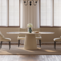 A dining room with a long oval table, upholstered chairs, a vase of white flowers, and stacked books, in front of windows with blinds.