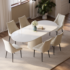 A white oval dining table with a fluted pedestal is surrounded by six cream upholstered chairs, with a vase of daisies and books on the tabletop.