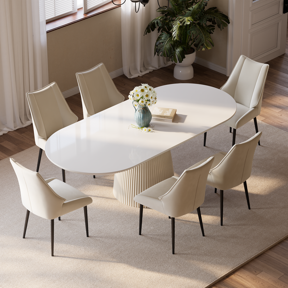 A white oval dining table with a fluted pedestal is surrounded by six cream upholstered chairs, with a vase of daisies and books on the tabletop.