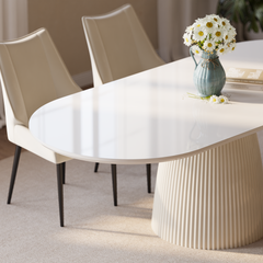 White dining table with a glossy rounded top and fluted pedestal base, paired with cream chairs with black legs, with white daisies in a blue pitcher and stacked books on top.