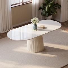 White oval table with a vertically fluted pedestal base, topped with a blue vase of white flowers and stacked books, on a beige rug beside a curtained window.