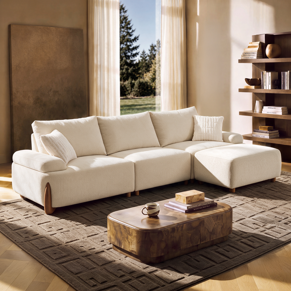 Cream sectional sofa with a right-side chaise and two throw pillows in a sunlit room with a patterned rug, low brown coffee table, open shelving and sheer curtains.