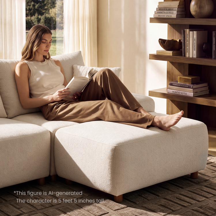 A woman reads a book while reclining barefoot on a cream-colored sofa with her legs stretched across an ottoman, beside a sunlit window and shelves holding books and ornaments.