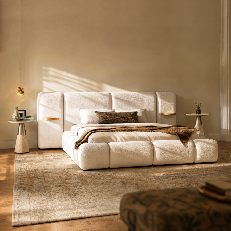 A cream upholstered bed with a tall segmented headboard sits in a warmly lit bedroom, with white pillows, a brown throw, bedside tables, and a large beige rug.