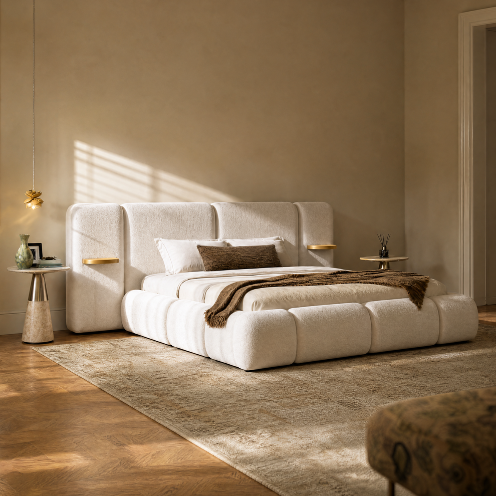 A white upholstered bed with a tall segmented headboard, white pillows, and a brown throw sits on a textured rug in a warmly lit bedroom.