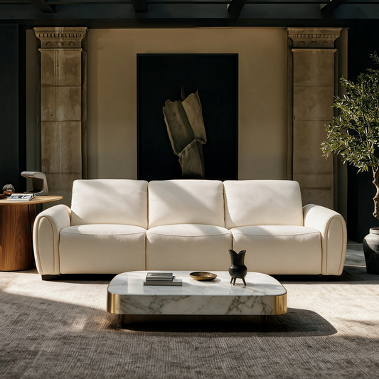 Cream three-seat sofa with rounded arms in a sunlit living room, with a low coffee table, dark framed abstract artwork, a side table and a potted tree.