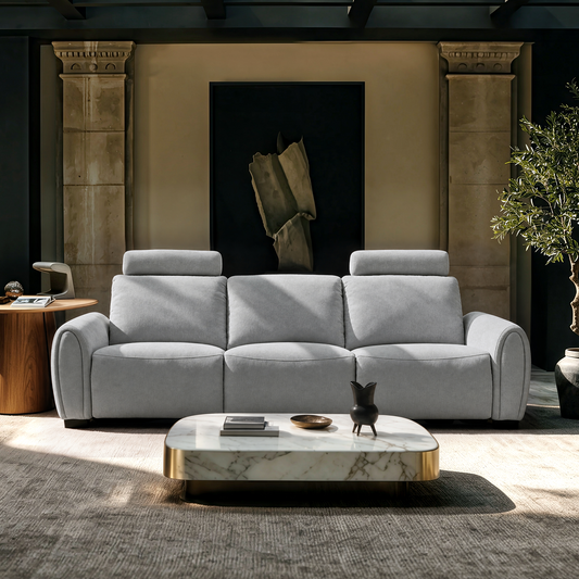 Light gray three-seat sofa with rounded arms and two raised headrests, set behind a low coffee table in a room with abstract wall art and a potted tree.
