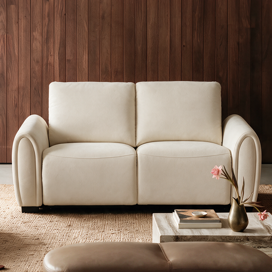 Cream two-seat sofa with rounded arms and two back cushions against a brown paneled wall, with a textured rug and a low table holding books and a vase of pink flowers.