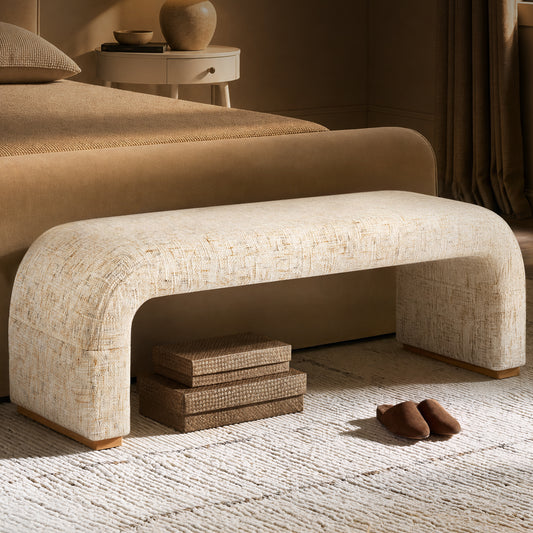 Cream upholstered bench with rounded corners and broad legs at the foot of a tan bed, with stacked woven boxes underneath and brown slippers on a textured rug.