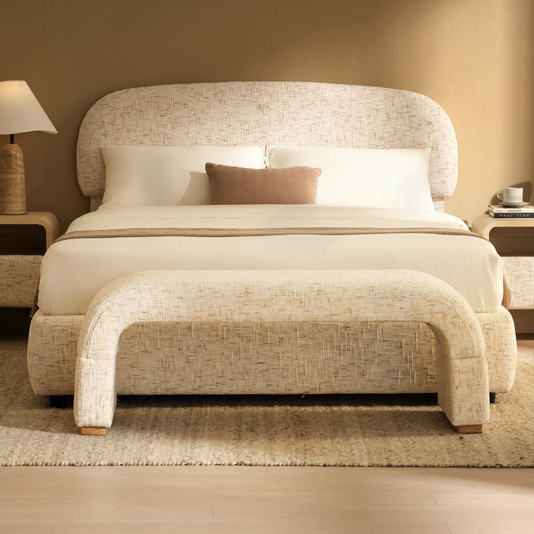 Cream upholstered bed with a wide rounded headboard, white bedding and a tan accent pillow, with a matching curved bench at the foot and bedside tables on either side.