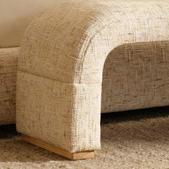 Close-up of a rounded furniture arm with textured cream upholstery, scattered darker threads, visible seams, and a light brown foot resting on a beige rug.