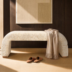 A cream upholstered bench stands against a dark wood-paneled wall, with a beige fringed throw draped over one end and brown slippers on the floor.