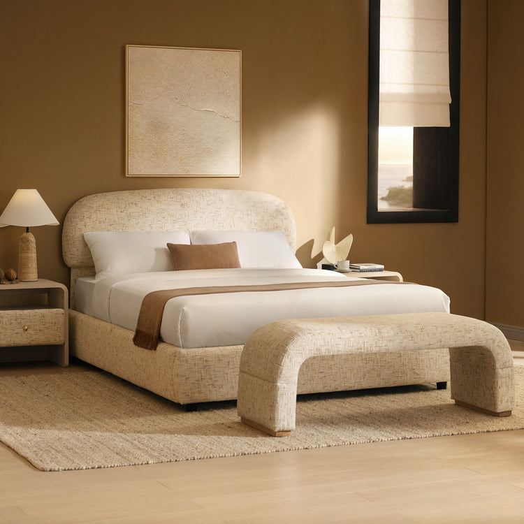 A cream upholstered bed with white bedding and a brown throw stands behind a matching curved bench in a warm-toned bedroom.