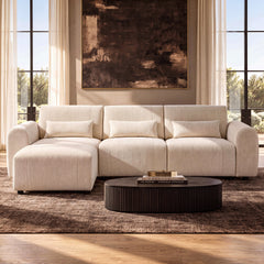 Cream sofa with rounded arms and three rectangular pillows, with a matching ottoman in front of the left seat and a dark oval coffee table beneath a large abstract painting.