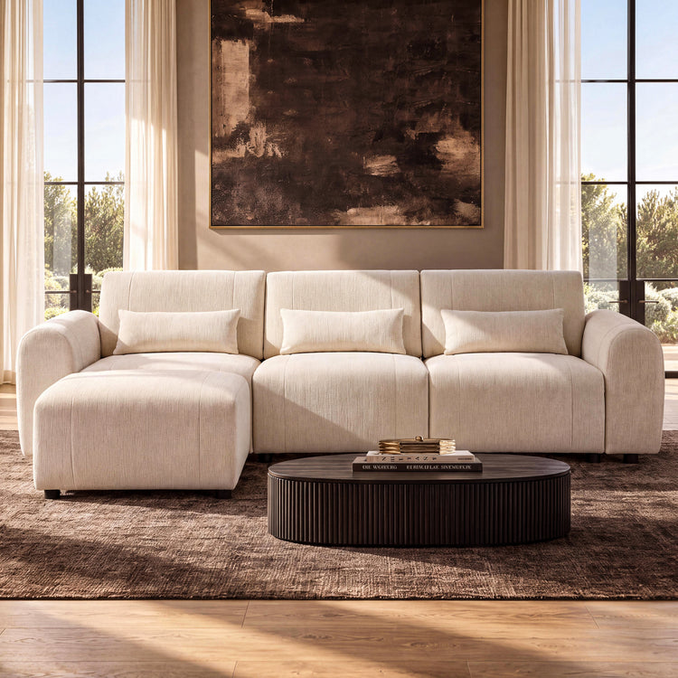 Cream sofa with rounded arms and three rectangular pillows, with a matching ottoman in front of the left seat and a dark oval coffee table beneath a large abstract painting.