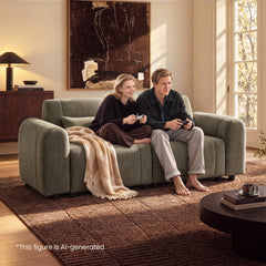 Two people holding game controllers sit on a green sofa with a beige fringed throw in a sunlit living room with abstract wall art and a brown rug.