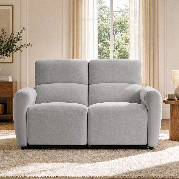 Front view of a light gray loveseat with two tall padded backs and rounded arms beside a living room window