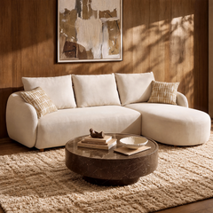 Cream sofa with a rounded chaise on the right and two patterned pillows, behind a round brown coffee table on a textured rug, with abstract artwork on a paneled wall.
