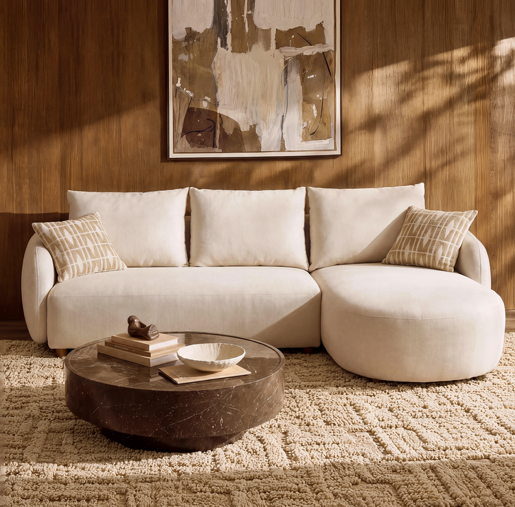 Cream sofa with a rounded chaise on the right and two patterned pillows, behind a round brown coffee table on a textured rug, beneath abstract artwork on a brown paneled wall.