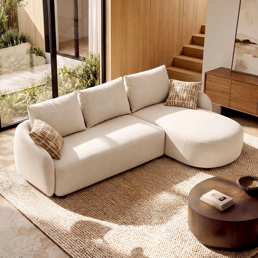 Cream sectional sofa with rounded arms, a right-side chaise, three back cushions and two patterned pillows, set on a textured rug beside tall windows in a sunlit living room.