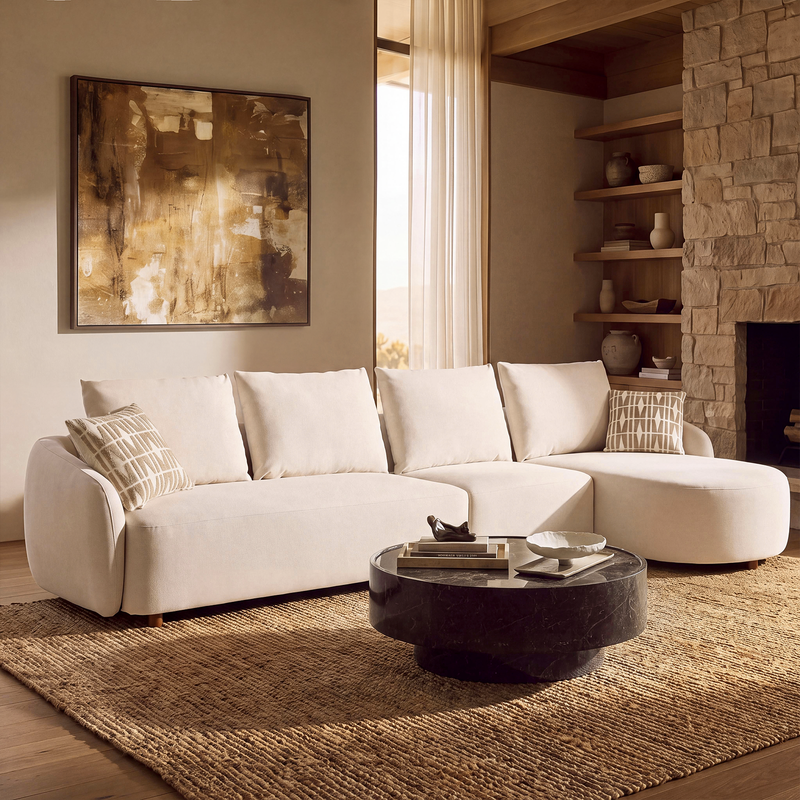 Cream sectional sofa with a rounded chaise and patterned cushions, facing a dark round coffee table on a textured tan rug, with abstract wall art, shelves and a fireplace behind.