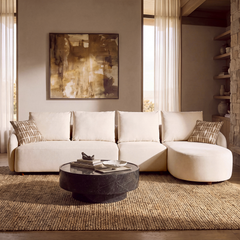 Cream sectional sofa with patterned pillows faces a round dark coffee table in a living room with abstract wall art, tall curtains, shelves, and a woven rug.