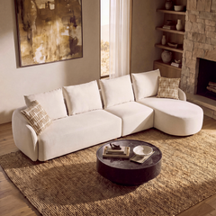 A cream sectional sofa with patterned cushions sits on a woven rug in a living room with a stone fireplace, shelves, and a round coffee table.