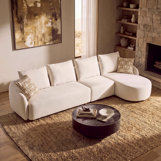 A cream sectional sofa with patterned cushions sits on a woven rug in a living room with a stone fireplace, shelves, and a round coffee table.